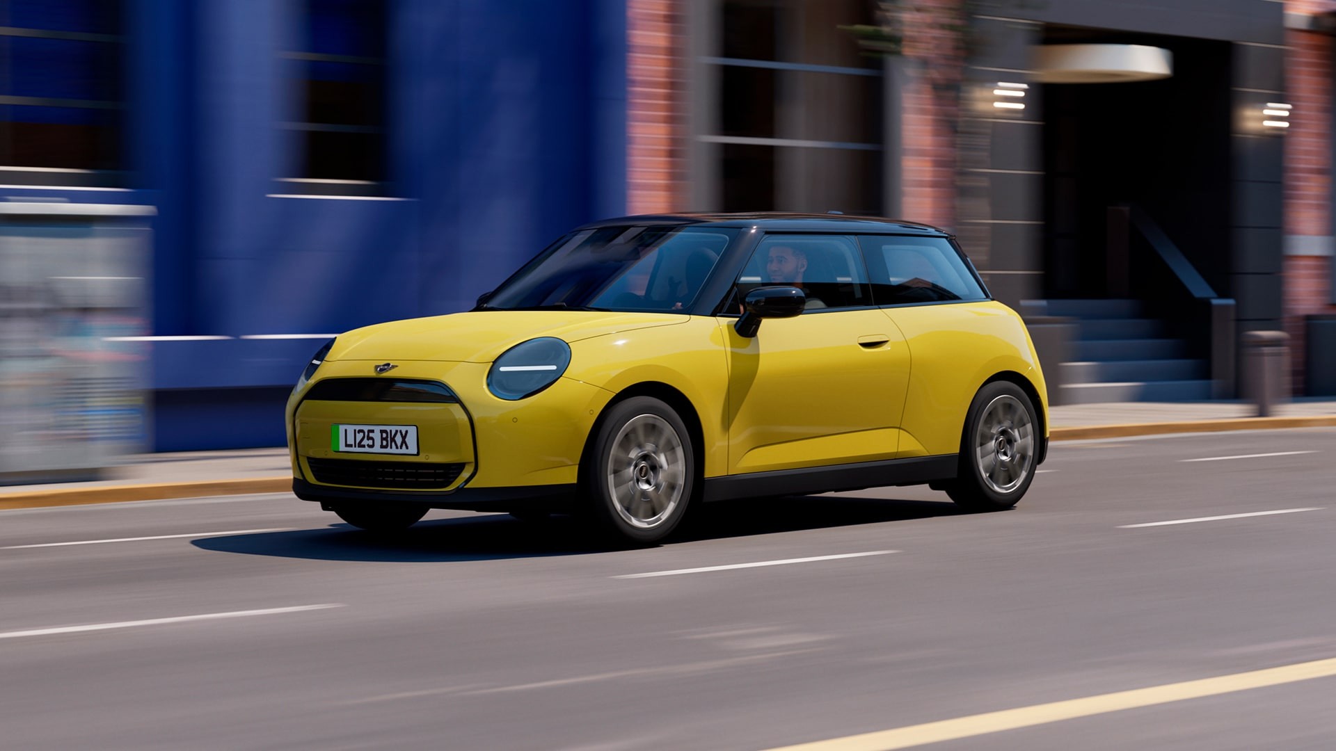 Yellow MINI Cooper electric hatchback driving on a city street with blue and brick buildings in the background, featuring black roof, circular LED headlights, and sleek modern design.